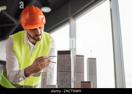 Bearded engineer in protective helmet working on building complex prototype project of residential or business district, measuring city model using compass for drawing at office with panoramic view Stock Photo