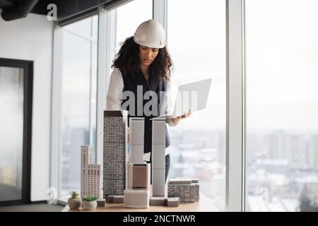 Focused woman in formal wear and white hardhat using digital laptop looking at design sustainable 3D megalopolis model on desk at open space office with panoramic windows with blurry cityscape. Stock Photo