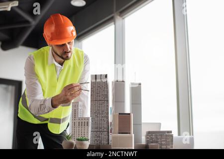 Bearded engineer in protective helmet working on building complex prototype project of residential or business district, measuring city model using compass for drawing at office with panoramic view Stock Photo