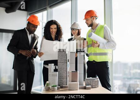 Focus on city model, multicultural workers reading blueprint. Architects, designers and engineers working on a building complex prototype project of residential or business district. Stock Photo