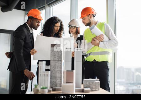 Multicultural workers in reflective vest and helmets with blueprint using city model. Architects designers and engineers works on building complex prototype project of residential or business district Stock Photo