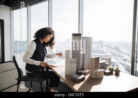 Curly female engineer working on building complex prototype project of residential or business district, measuring blueprint city model using compass at office with panoramic windows Stock Photo