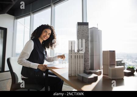 Curly female engineer working on building complex prototype project of residential or business district, measuring blueprint city model using compass at office with panoramic windows Stock Photo