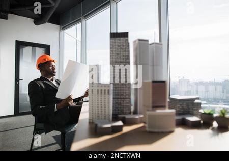 Office interior with panoramic city view. Handsome chief architect man in business suit and protective helmet holding blueprint sitting at desk working with city buildings model. Stock Photo