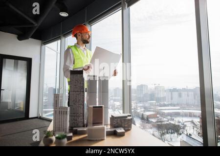 Engineer man in helmet and reflective vest holding blueprint standing near architecture design of city buildings, residential project maquette. Business man working on common construction project. Stock Photo