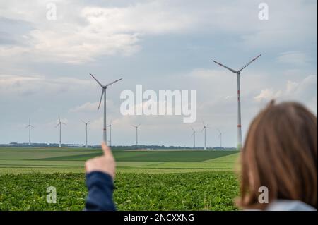 Woman with brown hair and blue jacket is pointing her finger on a wind turbine, wind park in agricultural field area, rear view Stock Photo