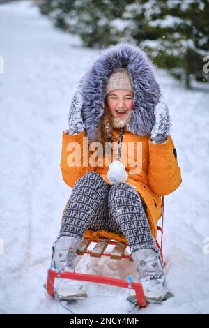 Kid girl enjoying snowy winter. Child sledding, sits on a sled and throws snow. Children play outdoors in snow. Kids sled in snowy park in winter. Win Stock Photo