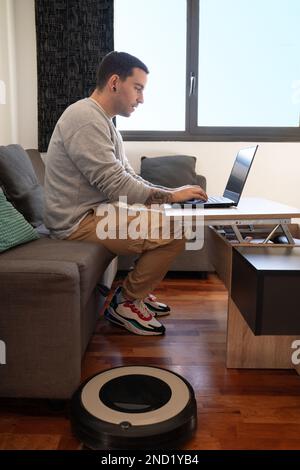 Side view full body of concentrated male freelancer in casual clothes sitting on sofa and working remotely via laptop near robot vacuum cleaner at hom Stock Photo