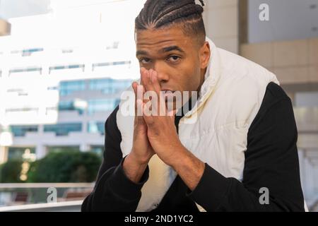 Serious young African American male with dreadlocks wearing casual clothes looking at camera while sitting against modern building with hands clasped Stock Photo