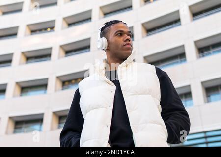 Low angle of calm young African American male in trendy outfit listening to music in wireless headphones while standing on city street Stock Photo