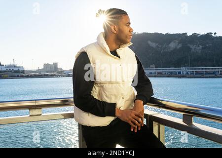 Confident young African American male with dreadlocks wearing casual outfit looking away while standing on embankment and leaning on railing against s Stock Photo