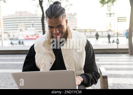 Smiling young African American male freelancer in casual clothes with dreadlocks sitting on bench and working remotely via laptop on street Stock Photo