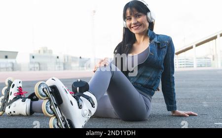 Positive young female in casual clothes and headphones smiling and looking at camera while listening to music and sitting on asphalt road in roller sk Stock Photo