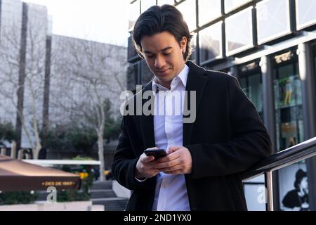 From below young male in classy outfit browsing on smartphone while standing near modern building and looking at screen Stock Photo
