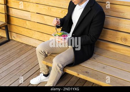 Cropped unrecognizable male in smart casual outfit sitting on wooden bench and having disposable bowl of takeaway healthy salad for lunch Stock Photo