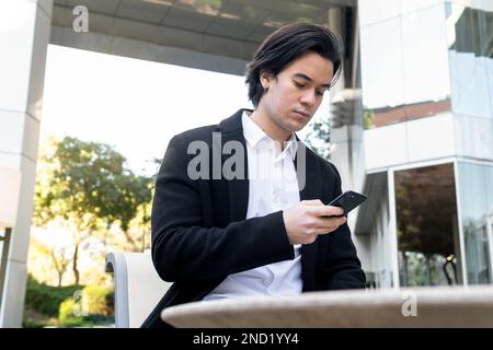 Concentrated young male in formal clothing sitting on chair and surfing internet via mobile phone at table in outdoor cafe Stock Photo