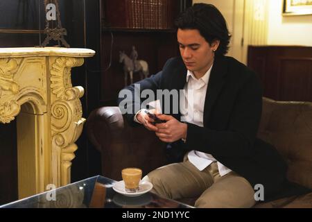 Focused young male in classy suit sitting on leather sofa and messaging via mobile phone near table with cup of coffee in cafe Stock Photo