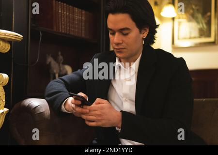 Focused young male in classy suit sitting on leather sofa and messaging via mobile phone near table with cup of coffee in cafe Stock Photo