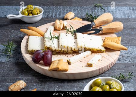 Appetizing cheese served on wooden table with ripe grapes and crackers decorated by rosemary sprigs near olives in bowls on table Stock Photo