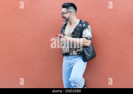 Cheerful bearded male hipster in summer outfit with sunglasses and bag looking away while standing against bright wall and browsing smartphone Stock Photo