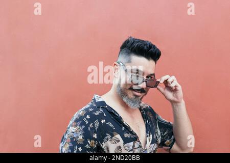 Side view of positive bearded male holding sunglasses and summer shirt smiling and looking at camera against bright wall Stock Photo