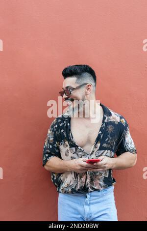 Positive bearded male hipster in summer shirt with sunglasses and mustache smiling brightly and looking away while browsing smartphone against red wal Stock Photo
