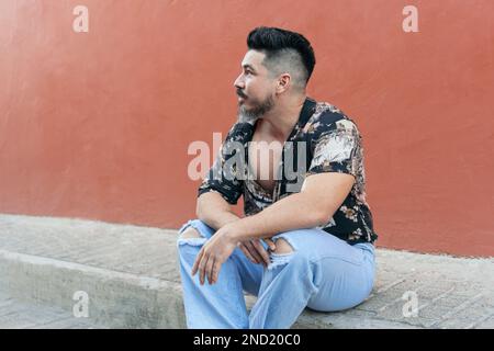 Thoughtful bearded male in casual clothes looking away while sitting on pavement against bright wall Stock Photo
