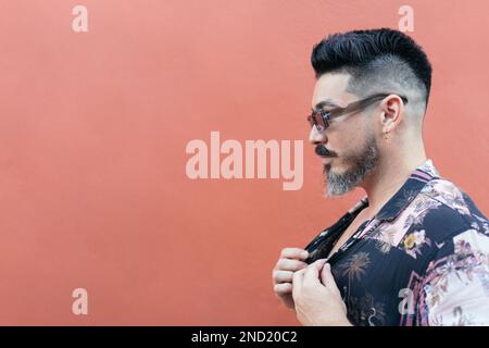 Side view of bearded male in sunglasses and summer shirt smiling and looking away against bright wall Stock Photo