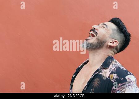 Side view of cheerful bearded male hipster in summer shirt laughing and looking up against bright wall Stock Photo