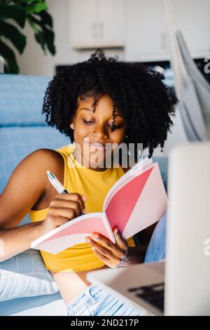 Delighted African American young female with Afro hair wearing casual outfit smiling and writing notes in planner while sitting near sofa at home Stock Photo