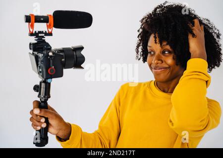 Confident smiling young African American curly haired female blogger in yellow sweatshirt standing against white wall and recording video on professio Stock Photo
