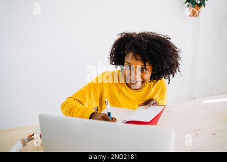 Positive young African American female student in casual outfit looking at screen of laptop while writing notes in copybook during exam preparation Stock Photo