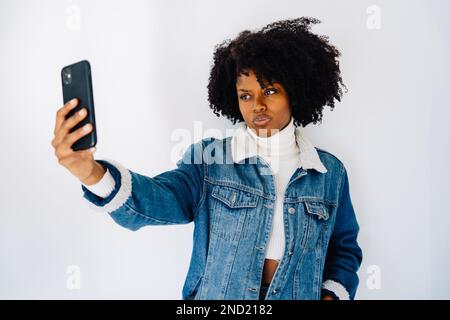Optimistic young African American female with Afro hairstyle in denim outfit while taking self portrait on smartphone against white background Stock Photo