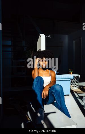 Pensive young African American curly haired female in casual outfit sitting on white seat and looking at camera while leaning on hand in daylight Stock Photo