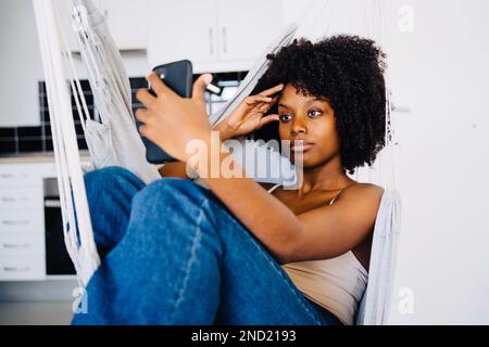 Emotionless young African American female in casual clothes with curly hair taking selfie on smartphone during rest on hammock Stock Photo