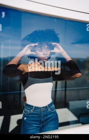 Through glass view of serious young African American female with Afro hairstyle touching hair while looking at window in daytime Stock Photo