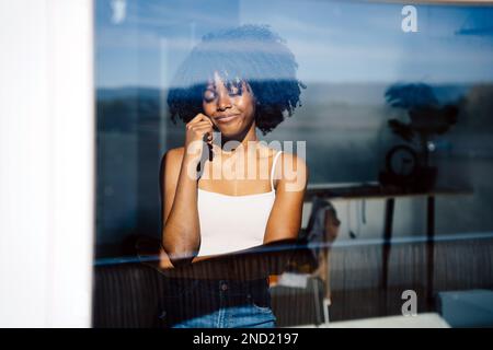 Through glass view of smiling young African American female with Afro hairstyle with eyes closed standing near windows in daytime Stock Photo