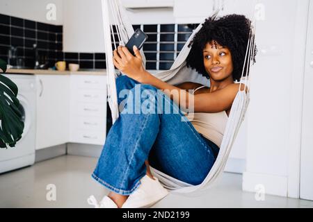 Cheerful young African American female in casual clothes with curly hair smiling taking selfie on smartphone during rest on hammock Stock Photo