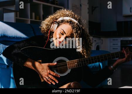 Cheerful young curly haired female musician in headphones playing acoustic guitar while sitting on floor near sofa and looking down Stock Photo