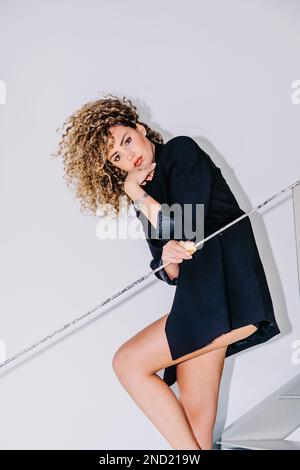 Confident young curly haired female in stylish black outfit looking at camera while leaning on railing against white wall Stock Photo