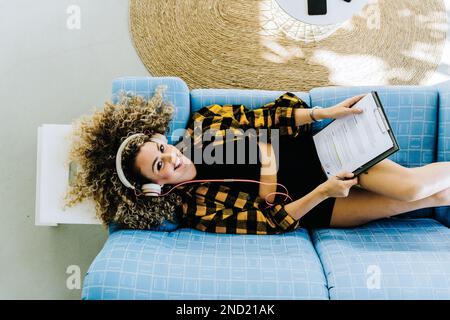 Top view of young smiling female in casual clothes lying on blue couch with clipboard while listening to music on headphones looking at camera Stock Photo
