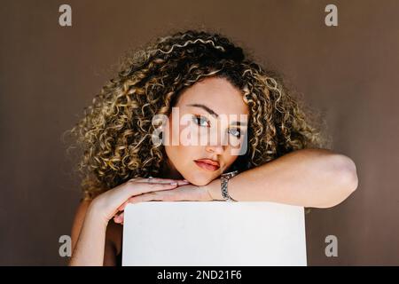 Young curly haired female with makeup and wristwatch looking at camera while leaning on cube in studio against brown background Stock Photo