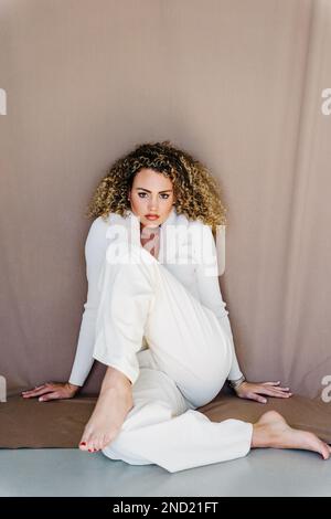 Full body high angle of young curly haired female in white outfit sitting with foot on knee against fabric brown backdrop looking at camera Stock Photo
