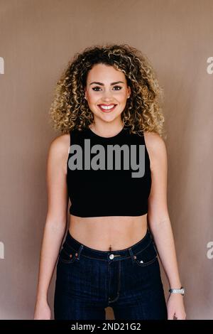 Cheerful young female with curly hair and wristwatch in black top and jeans standing in studio looking at camera Stock Photo