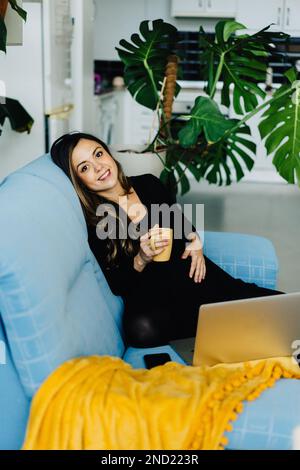 Smiling young woman carrying baby sitting on blue sofa and enjoying hot beverage while watching movie via computer Stock Photo