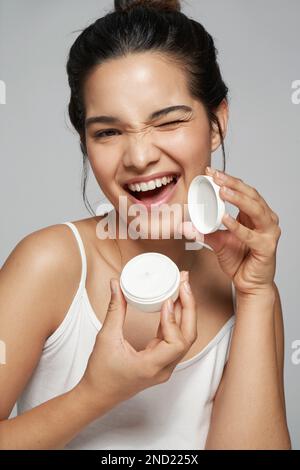 Delighted young female in white top smiling and looking at camera while opening bottle of cosmetic product Stock Photo