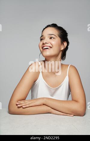 Positive young female in white top smiling and looking away while sitting against gray background with hands folded Stock Photo