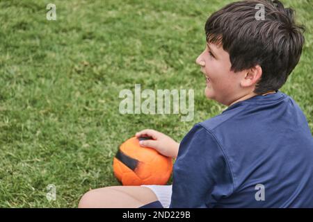 High angle of positive preteen boy in sportswear smiling and looking away while sitting on football field with ball Stock Photo