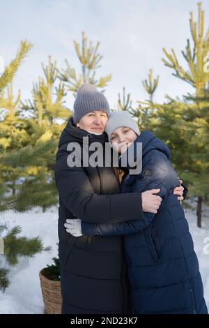 Positive mother and teen daughter in warm outwear and hats cuddling while looking at camera during weekend on winter day in forest Stock Photo