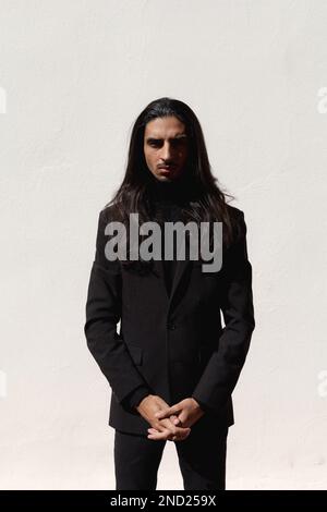 Confident young Hispanic male model with long dark hair wearing black classy suit standing against white background in studio and looking at camera Stock Photo